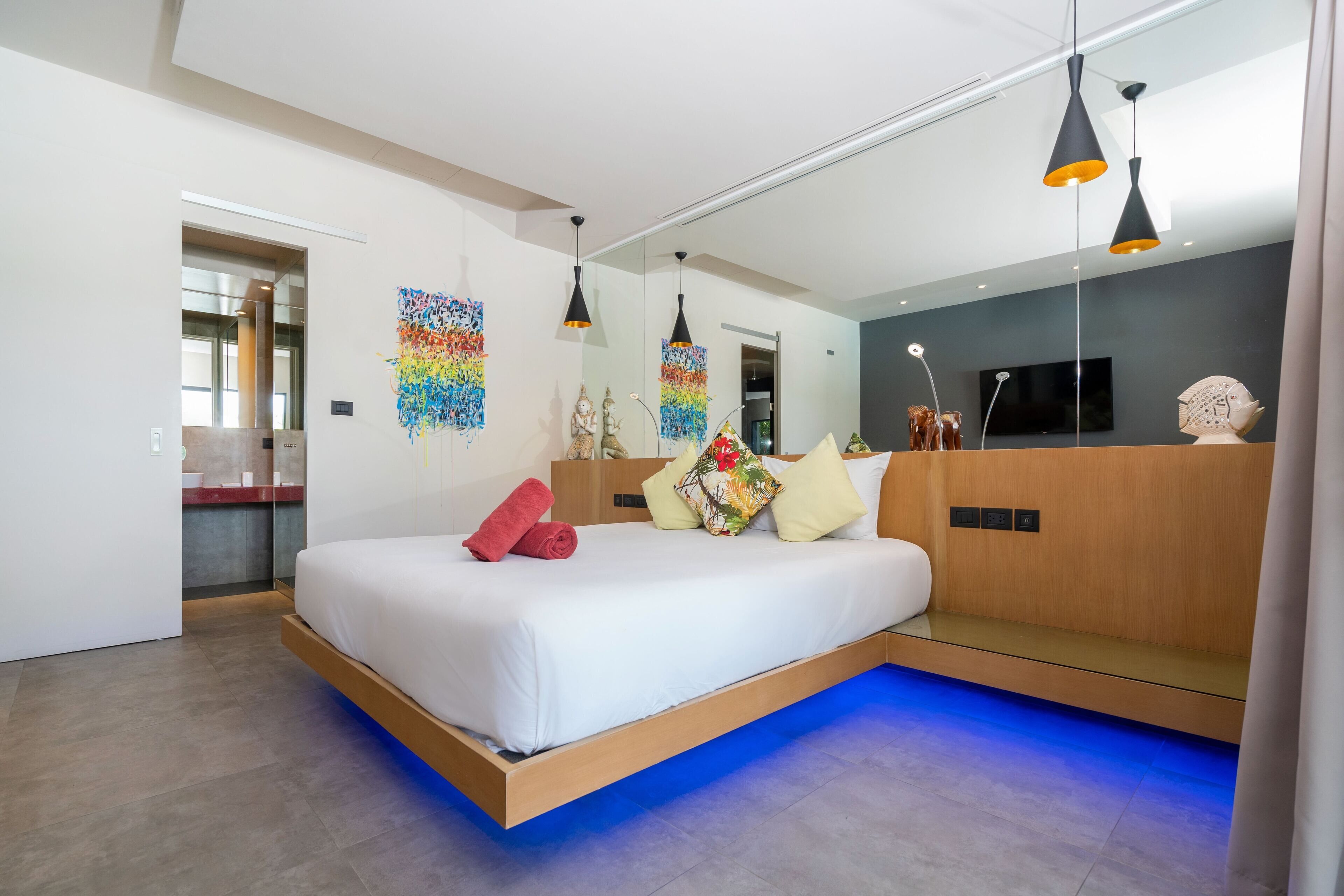 Een moderne hotelkamer met een groot bed, een houten hoofdbord en een ingebouwde bank. Er is een spiegel aan de muur, een flatscreen-tv en een plafondlamp.
