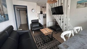 A modern living room with a black leather sofa, a wooden coffee table, and a patterned rug. There is a staircase with a TV mounted on it, leading to a kitchen area with white cabinets and a refrigerator. - 3 bedroom apartment in val d'arcomie (Val d'Arcomie)