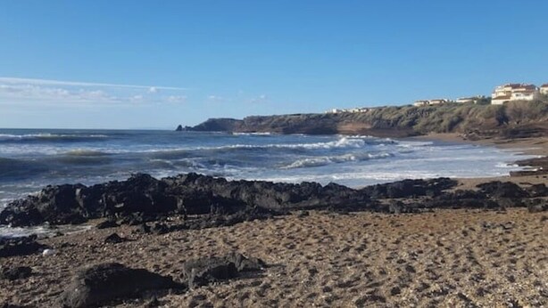 A rocky beach with waves crashing onto the shore, a clear sky, and buildings on a hillside in the background. - 3 bedroom apartment in val d'arcomie (Val d'Arcomie)
