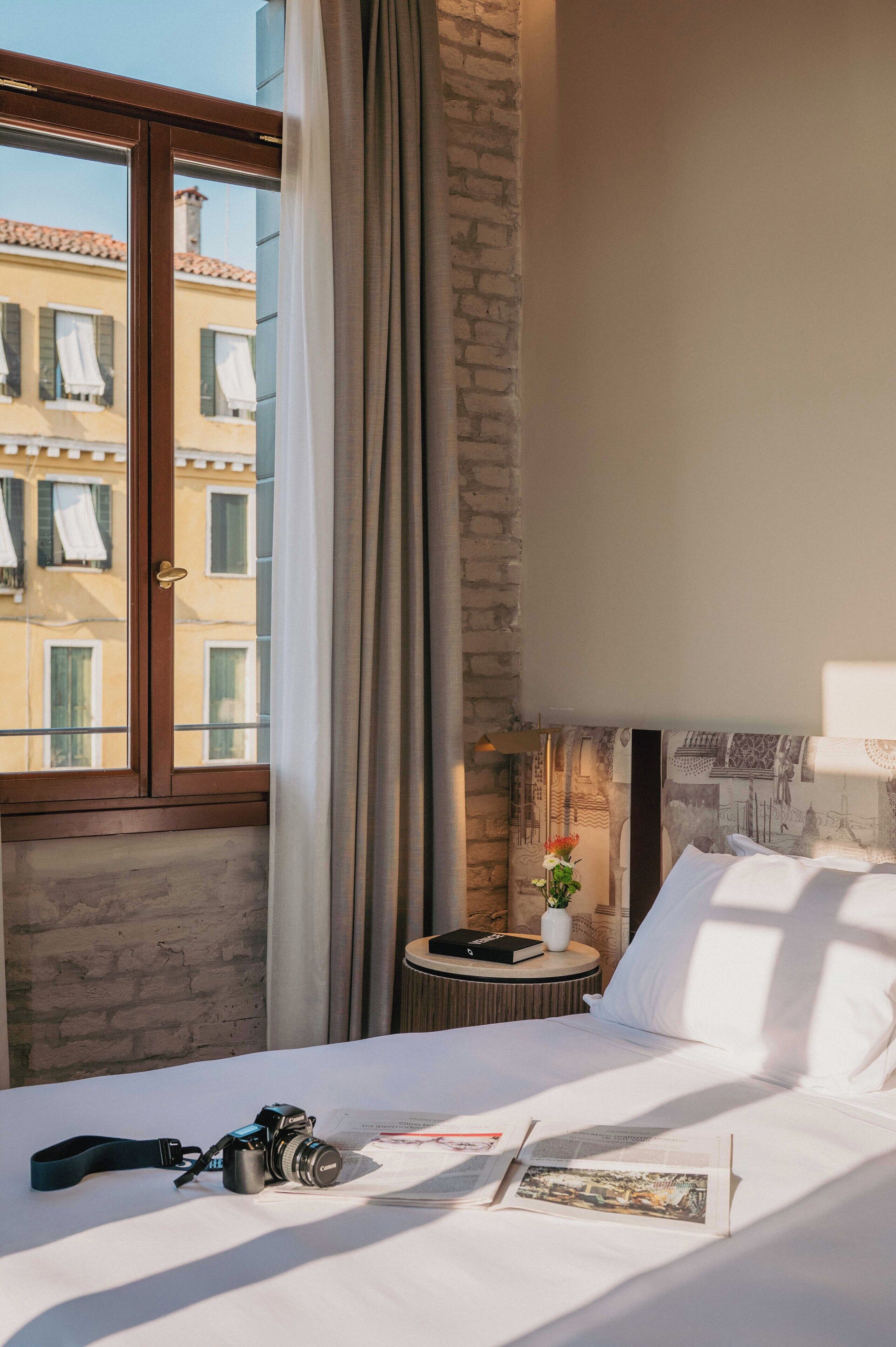 A hotel room with a bed, a camera, and a small table with a book and a vase of flowers.
