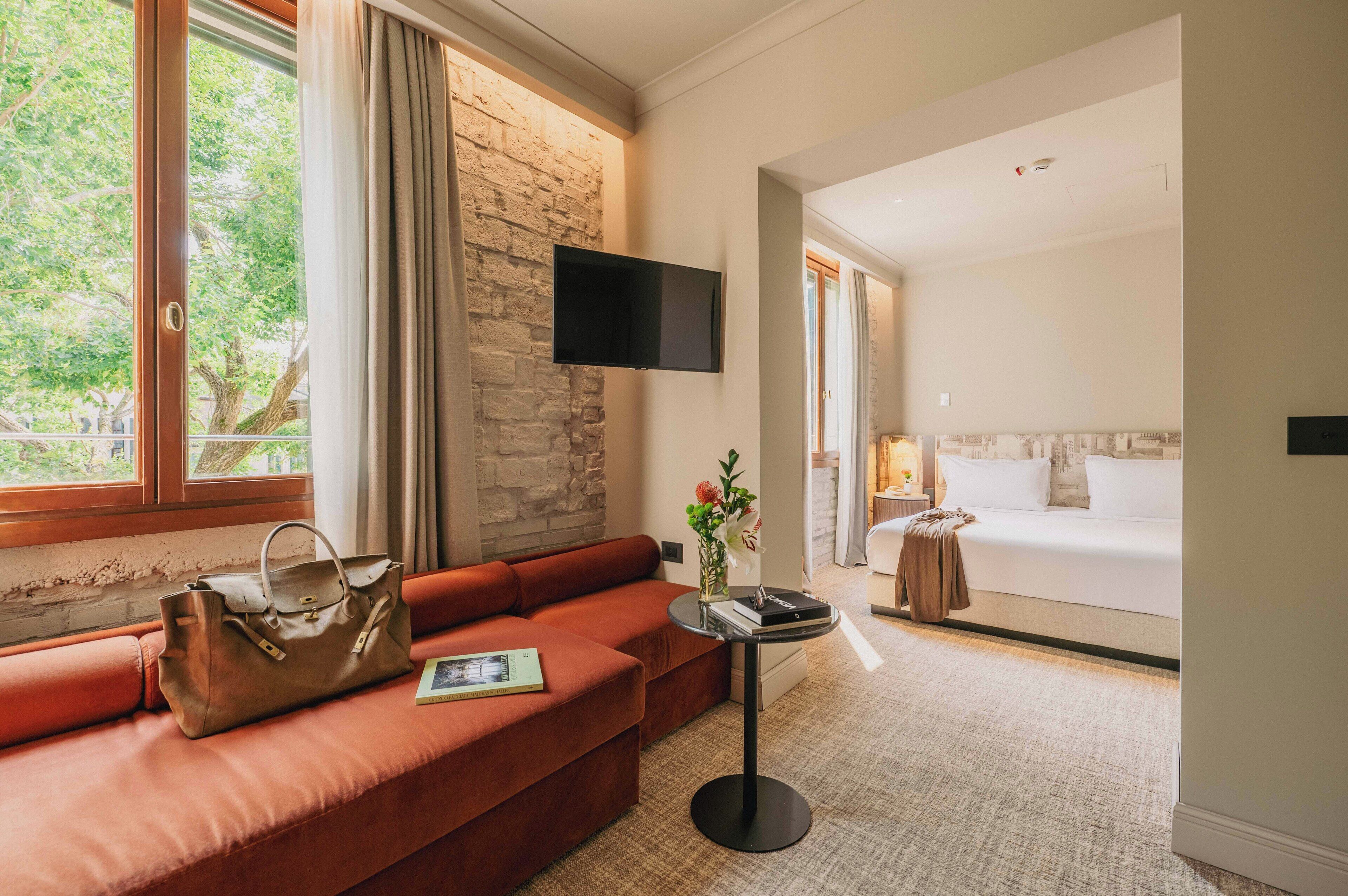 A hotel room with a red sofa, a flat-screen TV mounted on a stone wall, a bed with white bedding, and a small table with a vase of flowers.