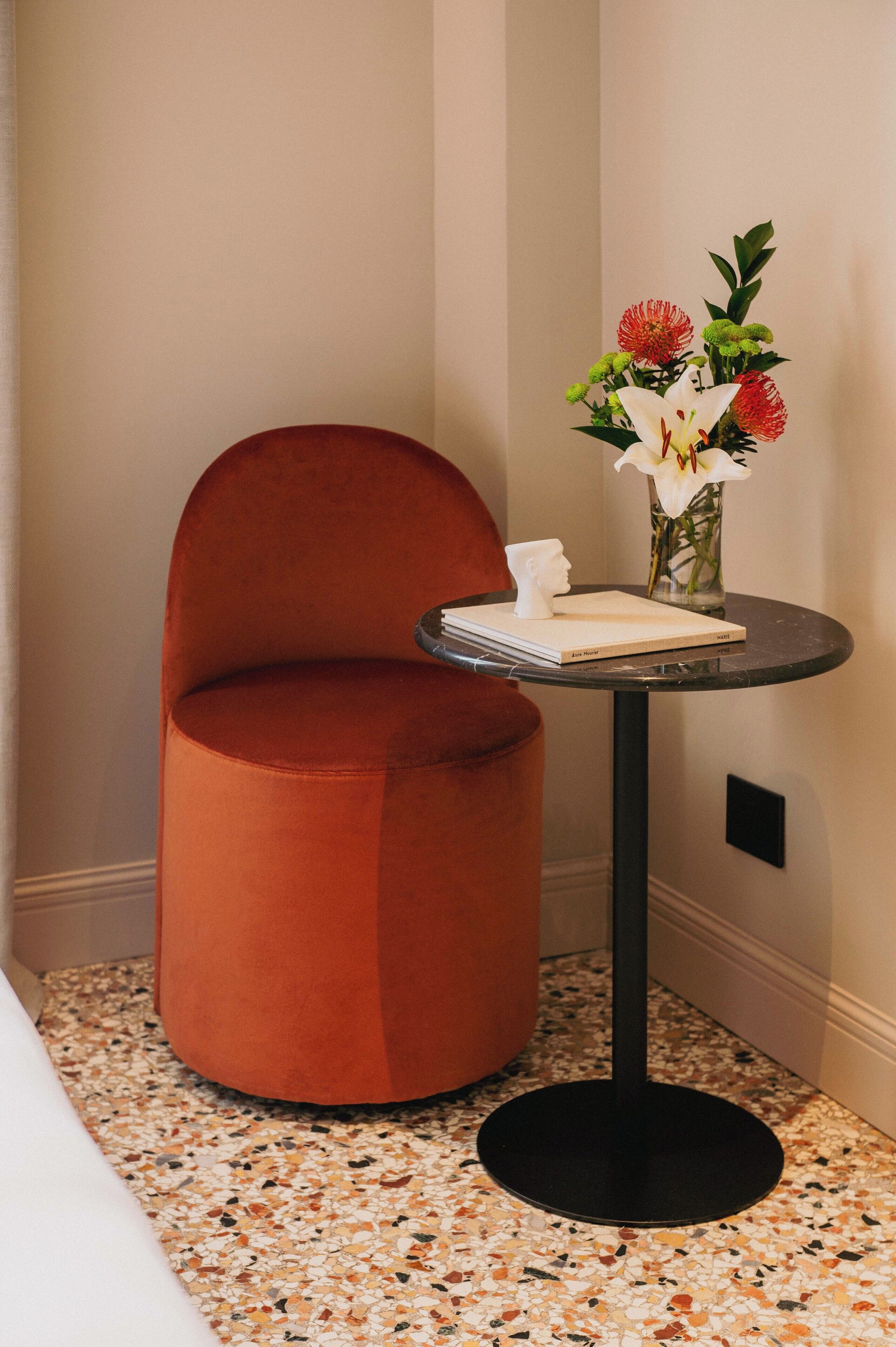 A corner room with a round orange chair, a small round table with a book and a vase of flowers, and a speckled floor.