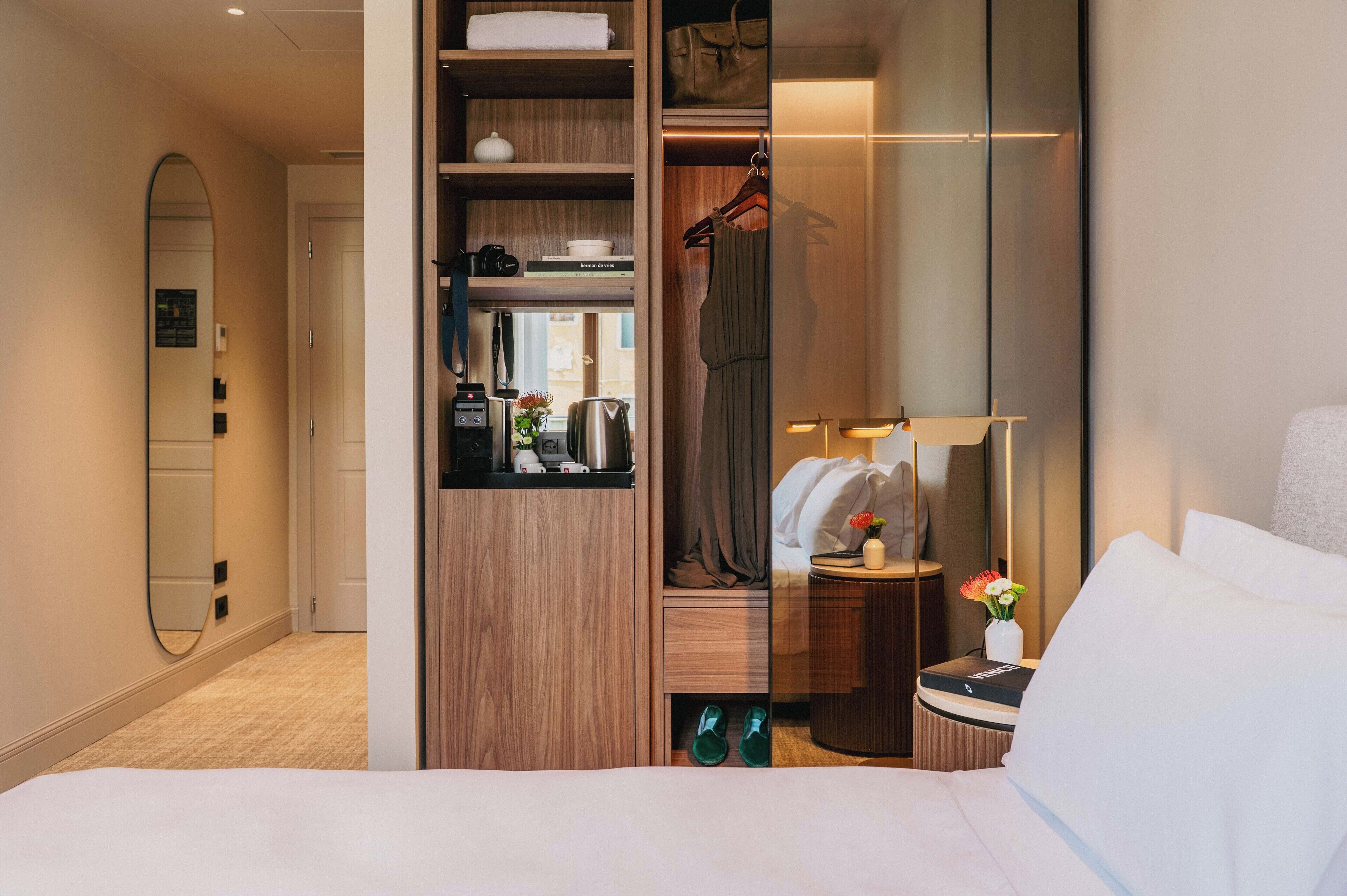 A hotel room with a bed, a mirror, a wardrobe, a vanity with a lamp, and a small table with a vase of flowers.