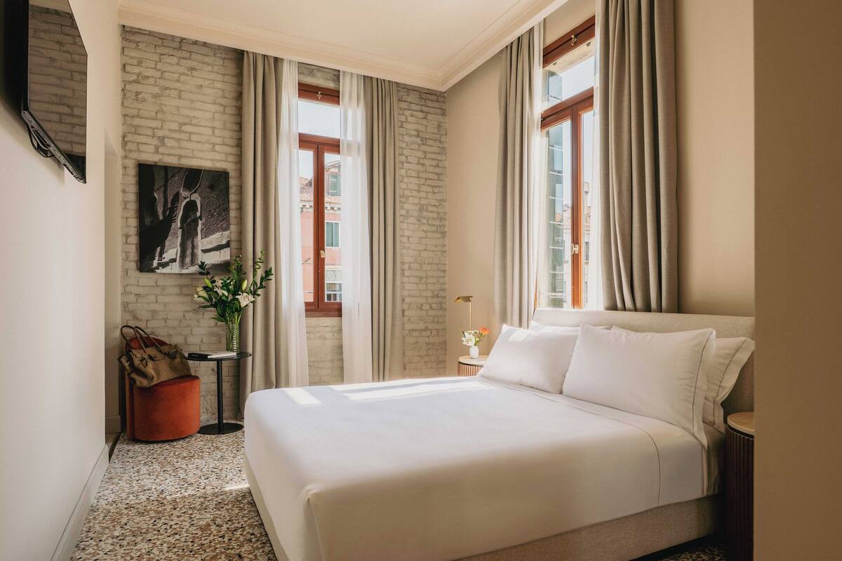 A hotel room with a large bed, a brick accent wall, a painting, a vase with flowers, and a view of buildings through the windows.