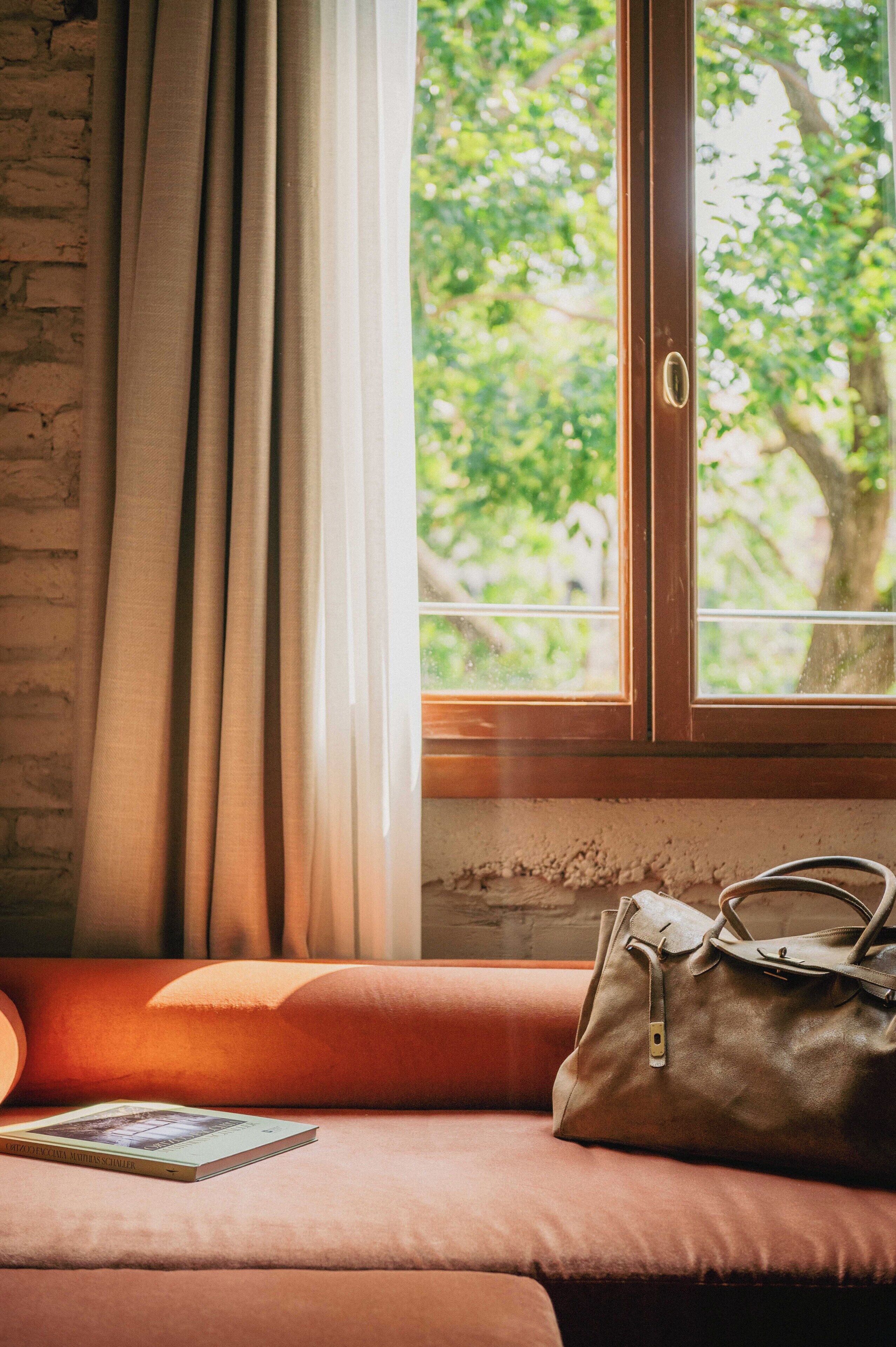 A room with a window, a brown leather bag, and a book on a cushioned seat.