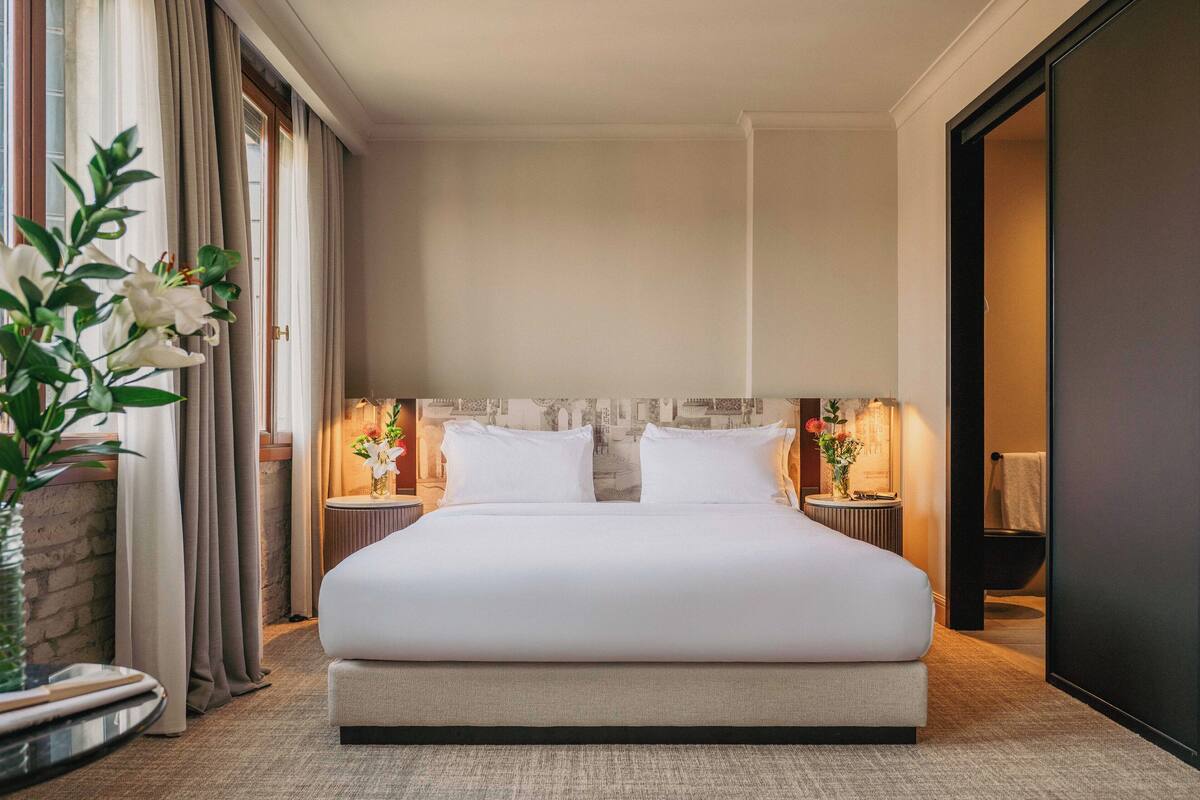 A spacious hotel room with a large bed, two bedside tables with lamps, a potted plant, and a view of the city through the window.