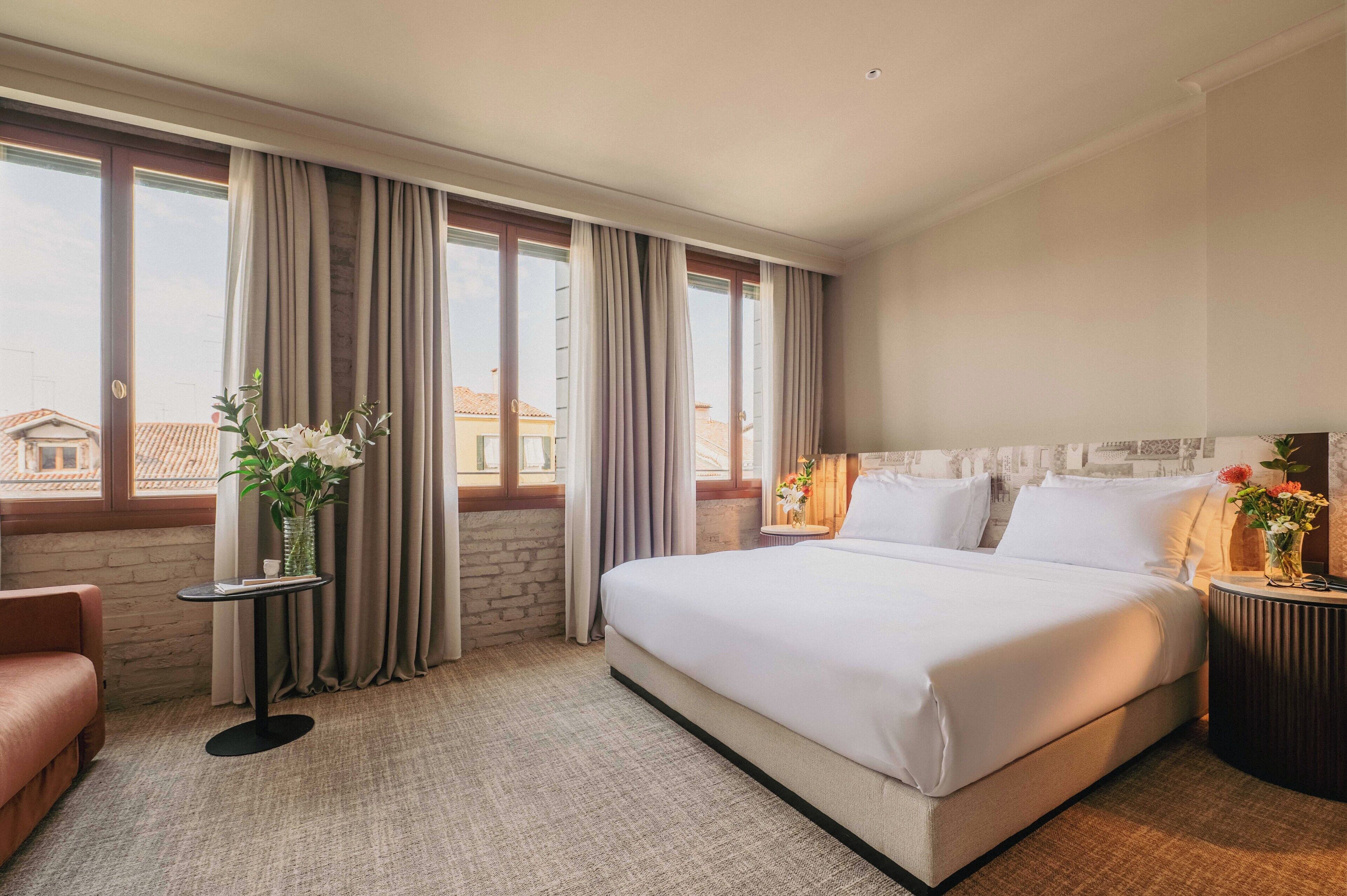 A hotel room with a large bed, a small table with a vase of flowers, and a view of buildings through large windows.