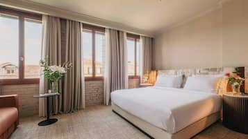 A hotel room with a large bed, a small table with a vase of flowers, and a view of buildings through large windows.