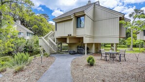 A two-story house with a covered patio, a staircase leading to the upper level, and a small garden area with a table and chairs. - Sunset Rentals | Night Heron 55 (Hilton Head Island)