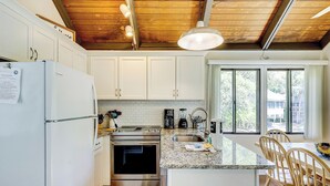 A kitchen with white cabinets, a stainless steel refrigerator, an oven, and a granite countertop. There is a window with a view of trees and a building outside. - Sunset Rentals | Night Heron 55 (Hilton Head Island)