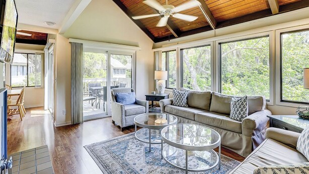 A spacious living room with a high ceiling, large windows, and a ceiling fan. - Sunset Rentals | Night Heron 55 (Hilton Head Island)