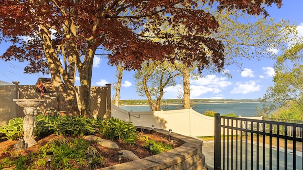 A landscaped garden with a stone wall, a decorative fountain, and a view of a body of water. - Coastal Comfort With Stunning Harbor Views 3 Bedroom Home (Plymouth)