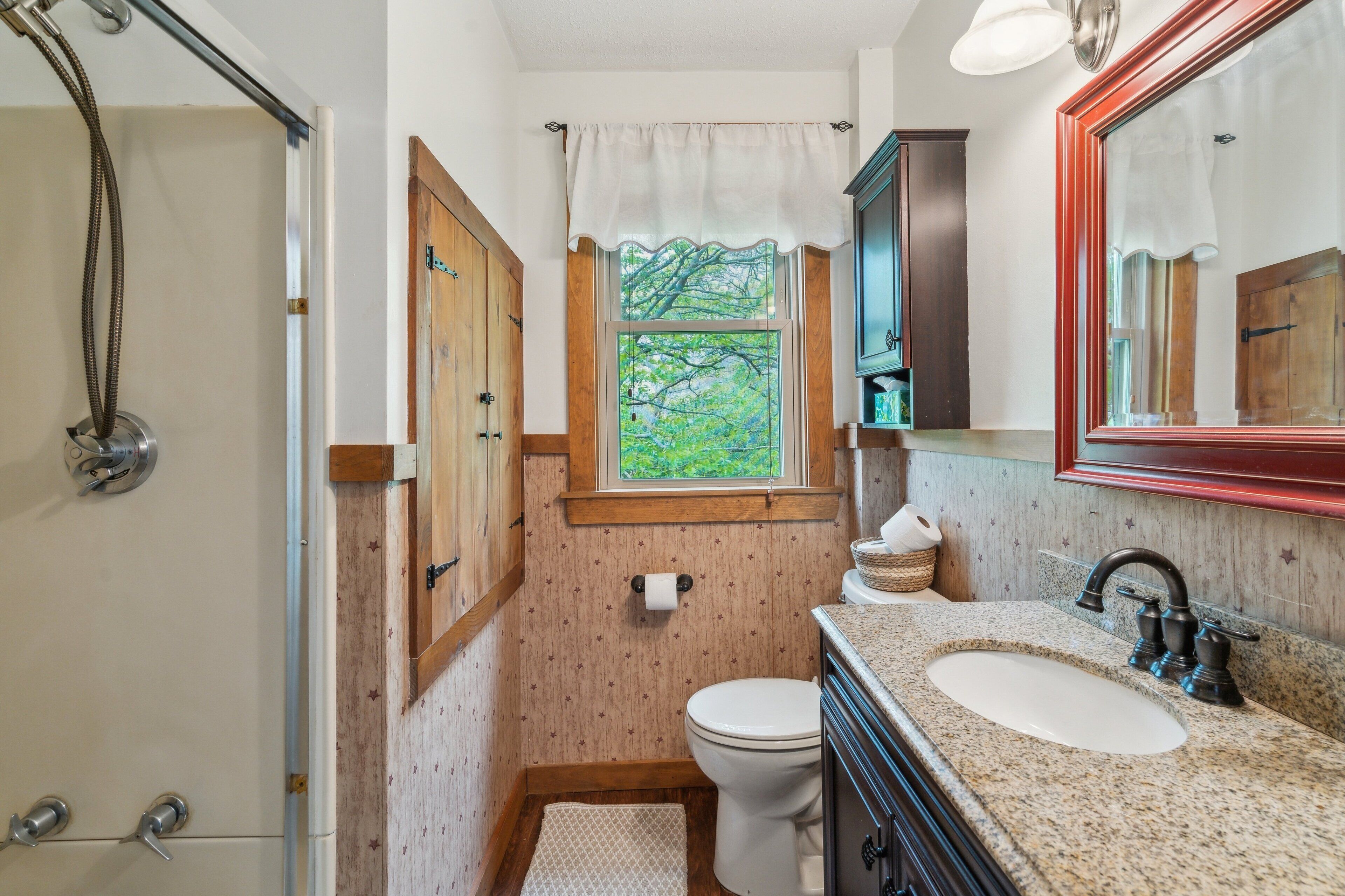 A bathroom with a shower, toilet, sink, and a window with a view of trees.