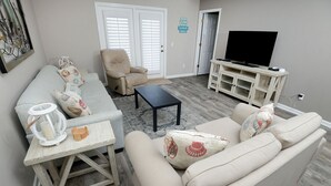 A living room with a sofa, armchair, coffee table, TV stand, and a door with a sign. - 225 Seahorse Way 3 Bedroom Home (Panama City Beach)