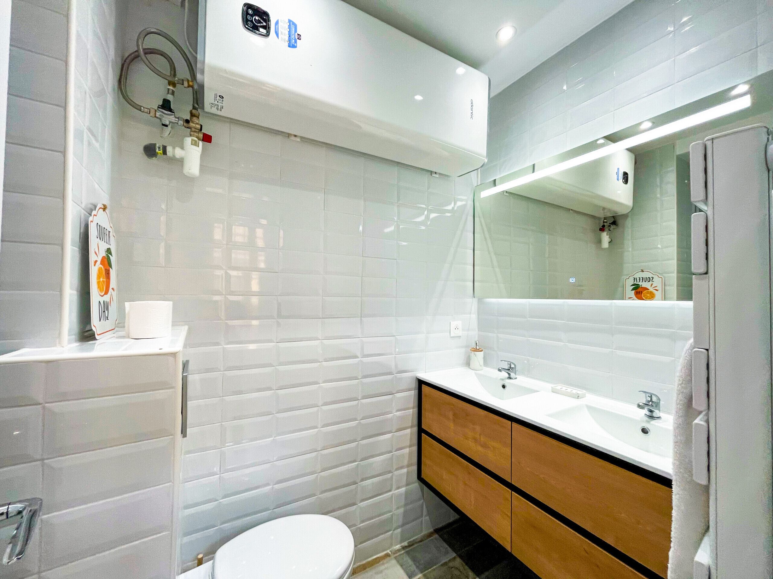 Ein modernes Badezimmer mit weißen Fliesen, einer weißen Toilette, einem hölzernen Waschtisch mit Spiegel und einem Handtuchhalter.
