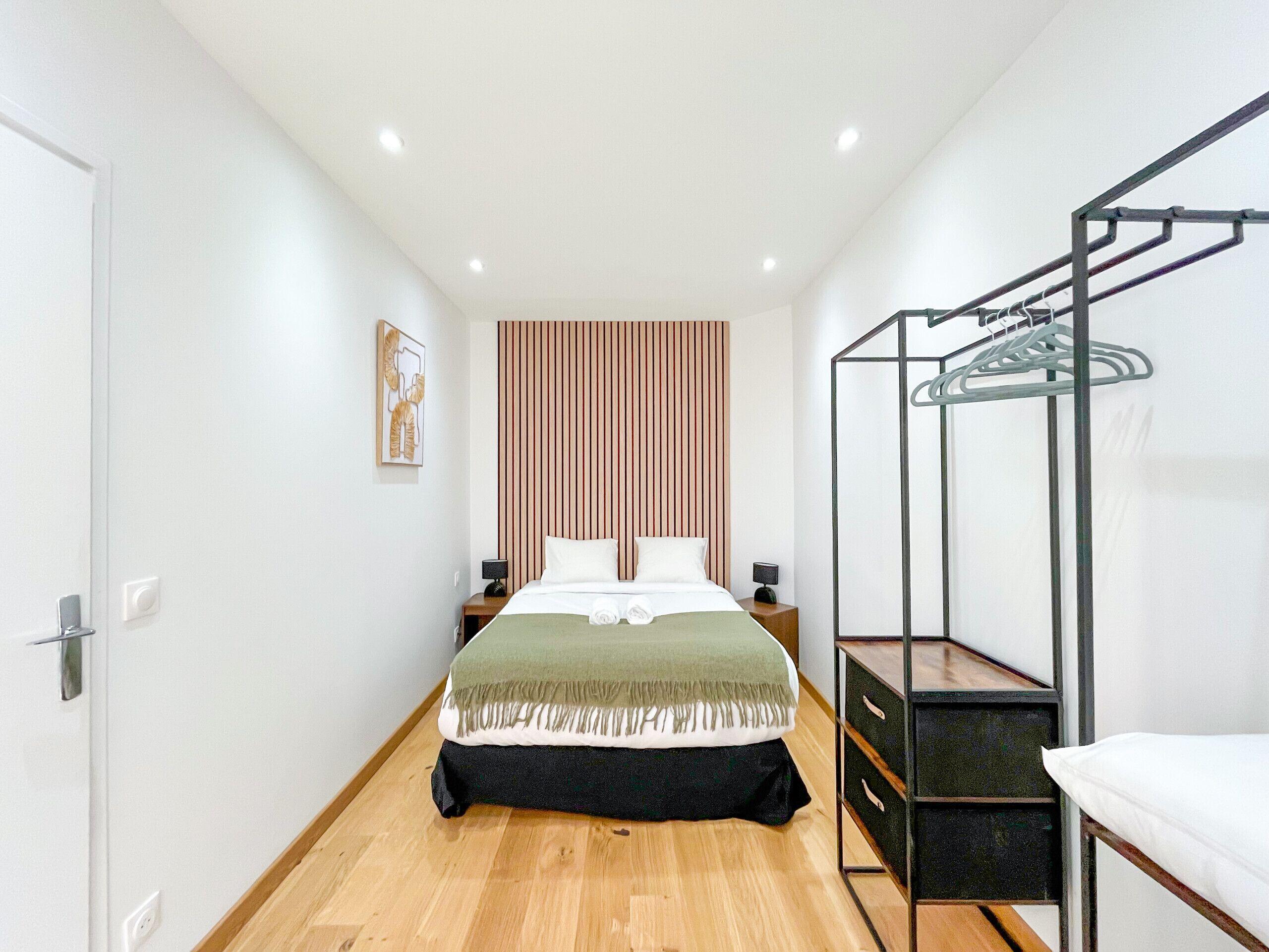 Una habitación de hotel moderna con suelo de madera, una cama con cubrecama verde y blanco, un panel de pared de madera, un armario metálico y un perchero de pared.