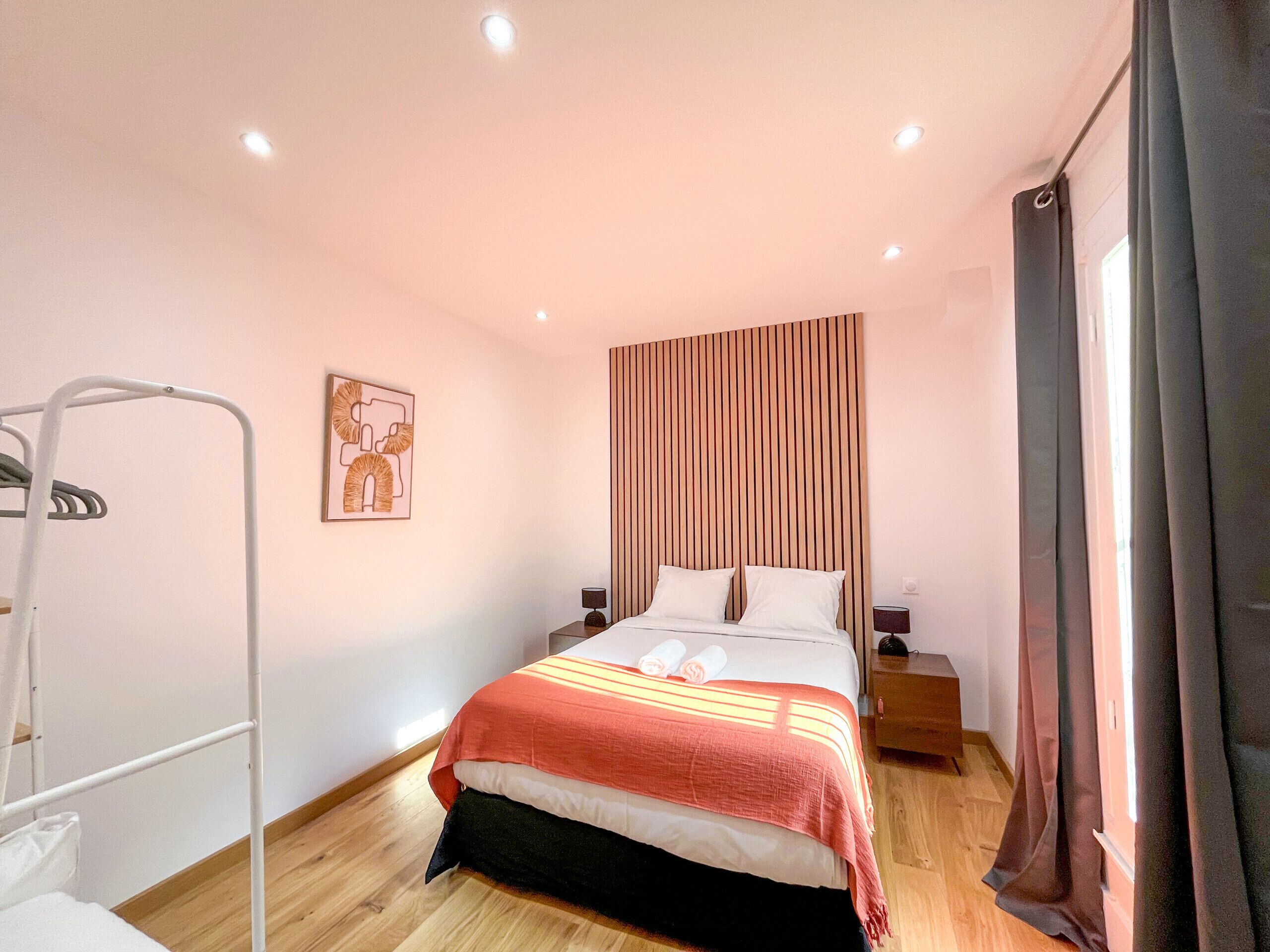 Un dormitorio con cama, mesita de noche, estante en la pared y ventana con cortinas.
