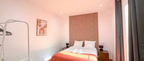 Un dormitorio con cama, mesita de noche, estante en la pared y ventana con cortinas.