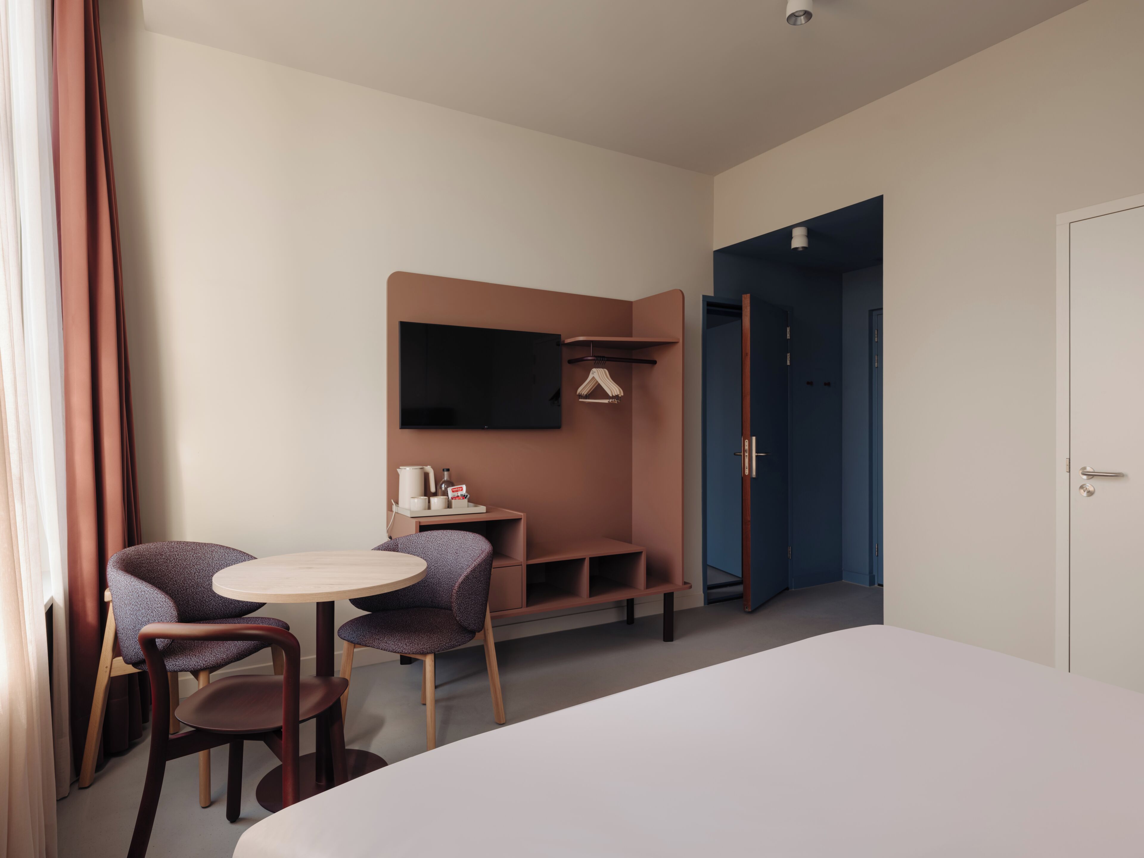 A hotel room with a bed, a desk with a chair, a TV mounted on the wall, and a wardrobe.