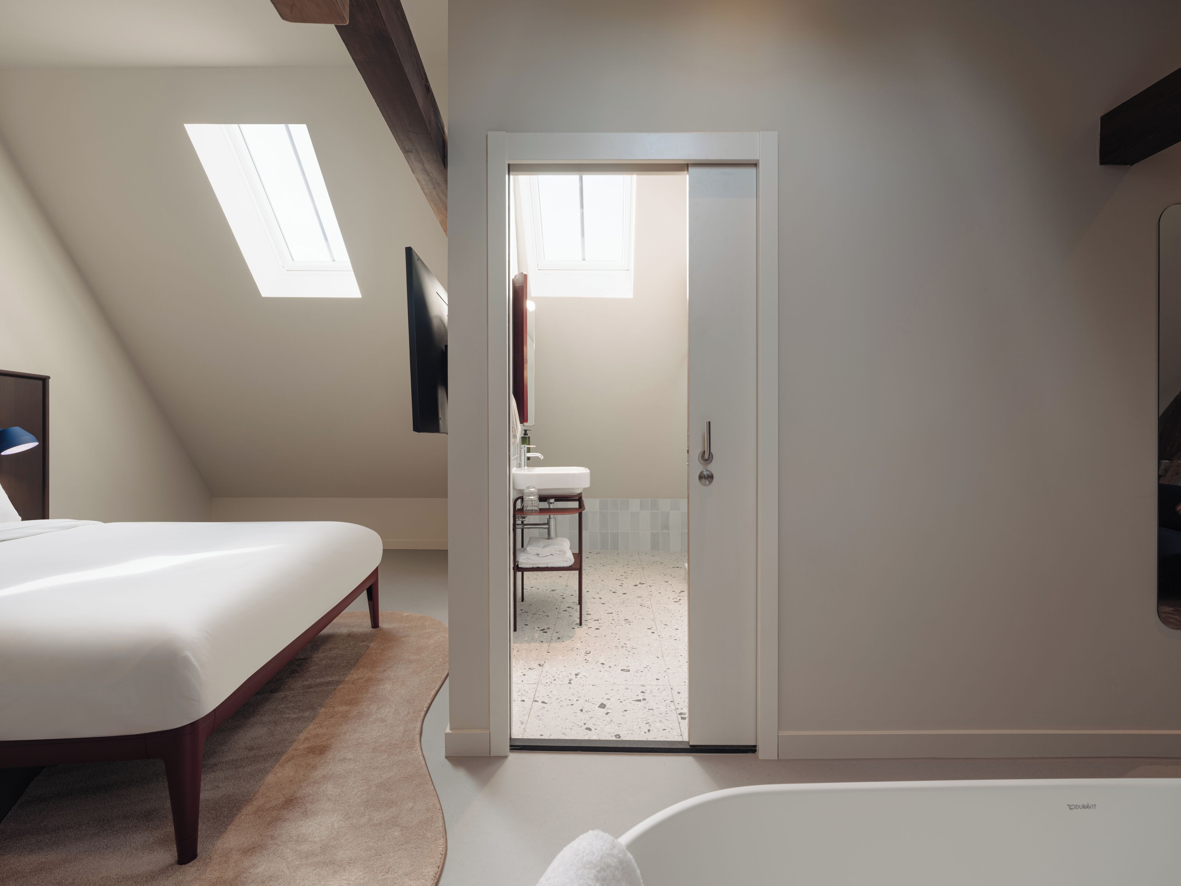 A bedroom with a bed, a desk, a chair, a window, and a door leading to a bathroom.