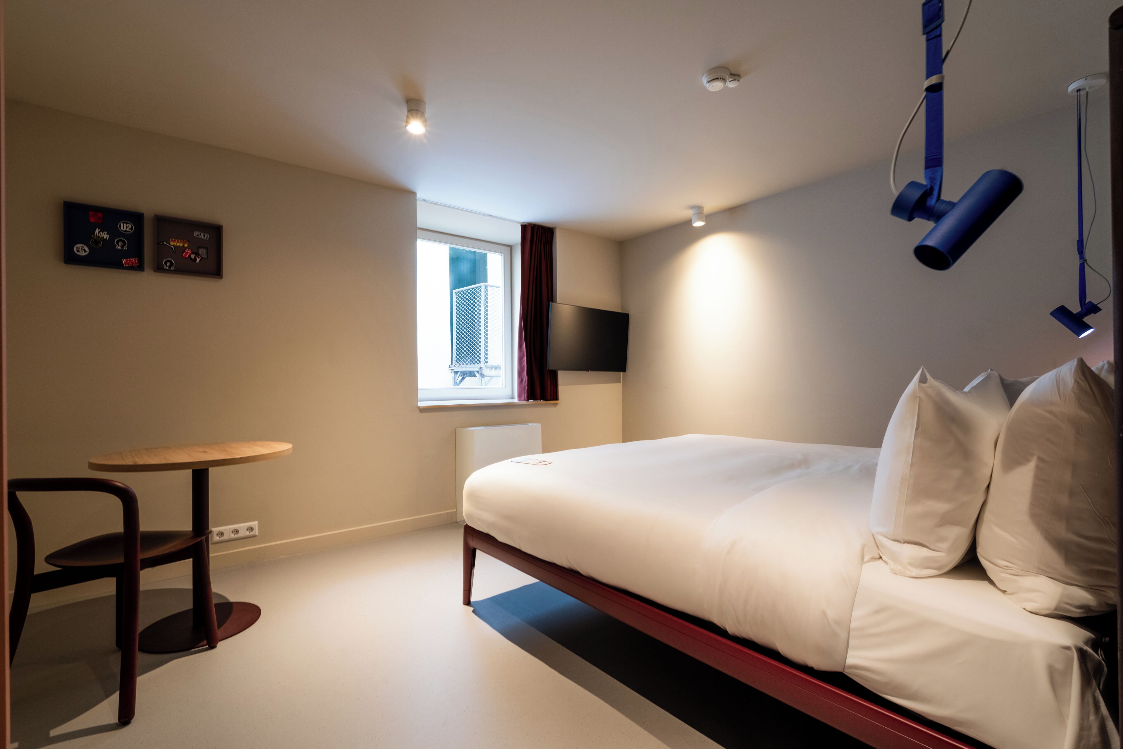 A modern bedroom with a bed, a small table, a chair, a window, and a flat-screen TV mounted on the wall.