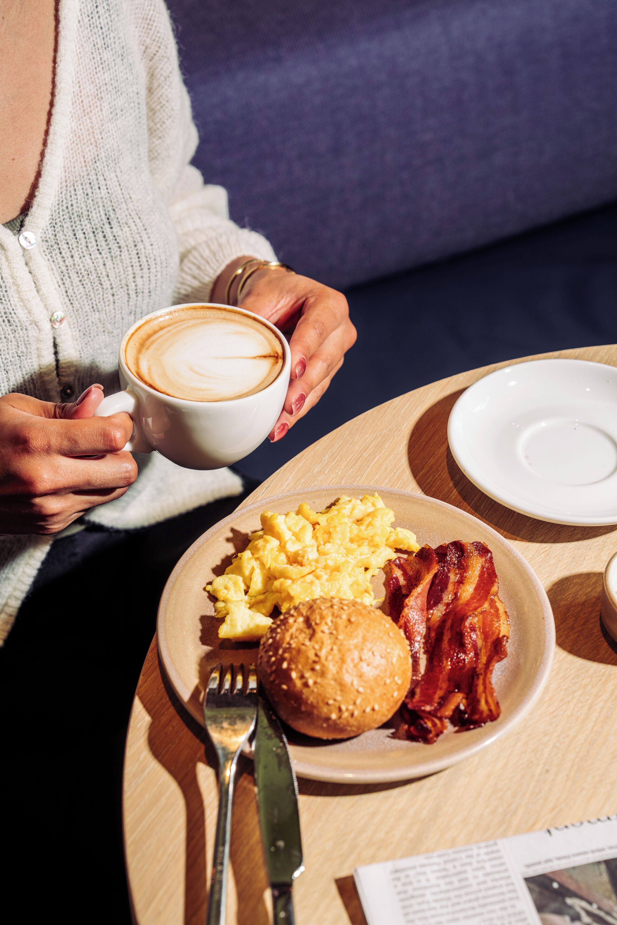A person holding a cup of coffee, sitting at a table with a plate of breakfast including scrambled eggs, bacon, and a hamburger.