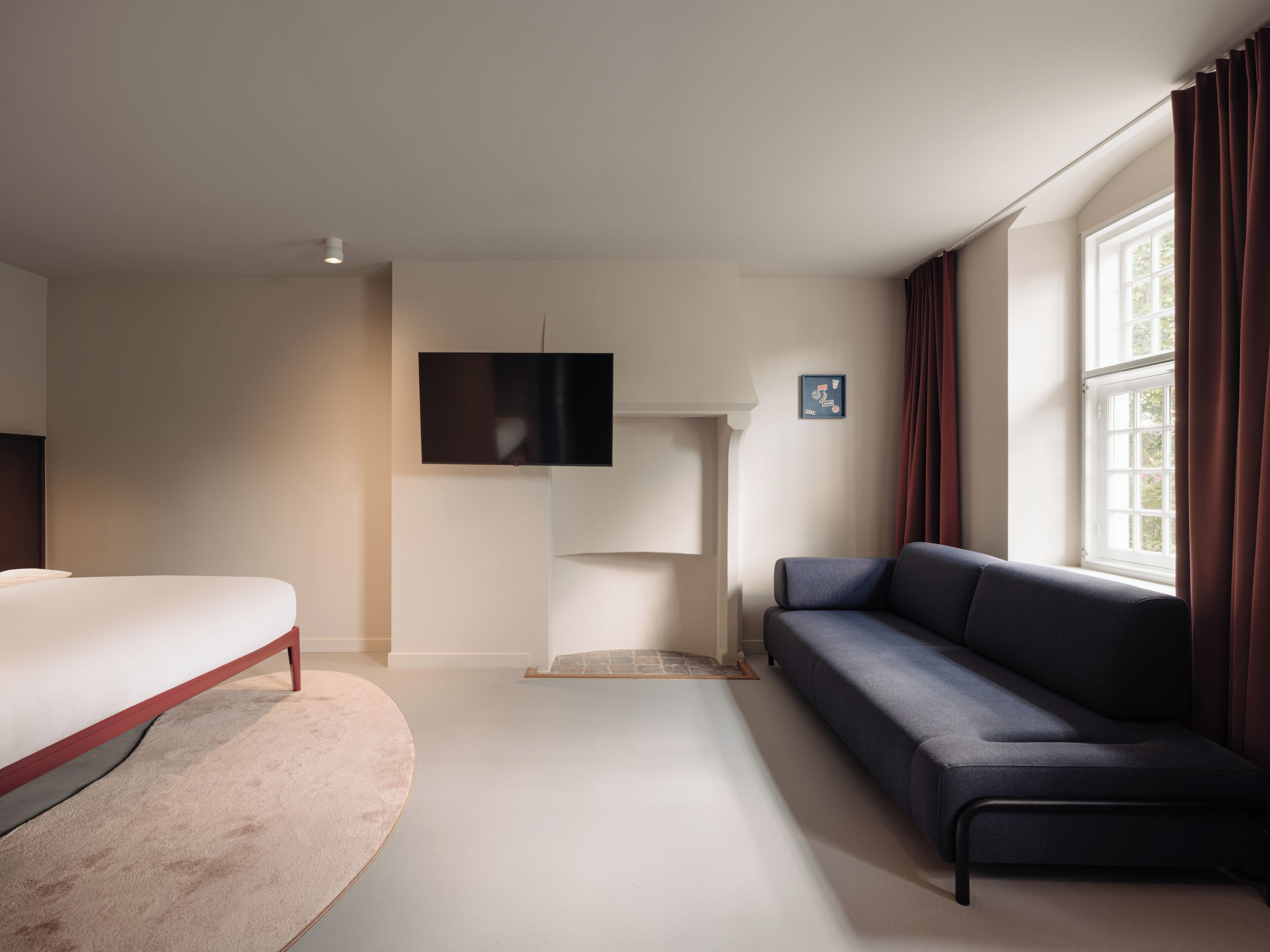 A modern hotel room with a bed, a sofa, a flat-screen TV mounted on the wall, and a large window with curtains.