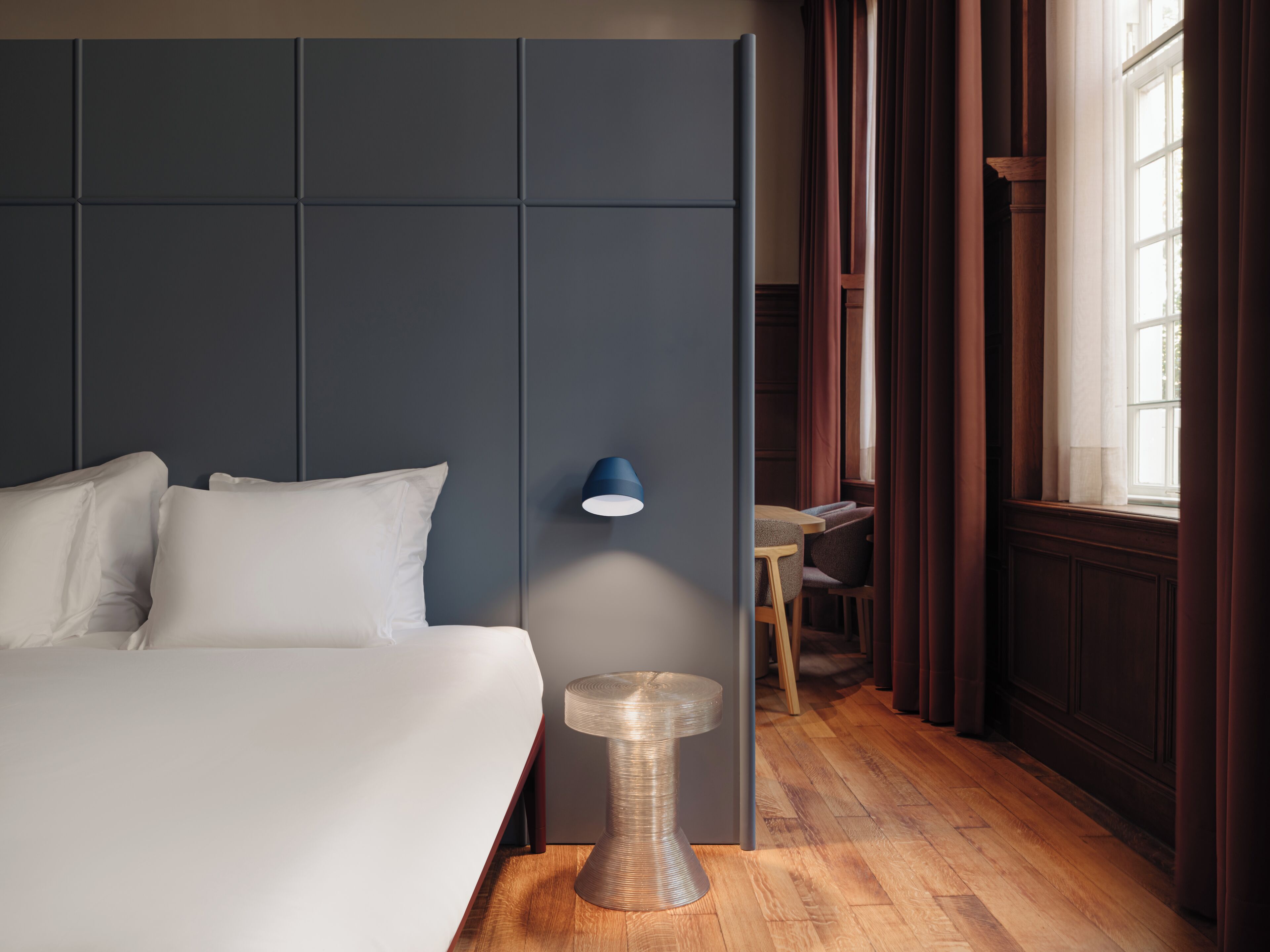 A modern hotel room with a large bed, a small desk, a lamp, and a view of the outdoors through a window.