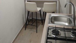 A compact kitchen with a gas stove, a sink, and two white chairs. There are wall-mounted decorations including a cactus painting and a 'HOME' sign. - Cozy two-room apartment in the heart of Netino Baroque (Noto)