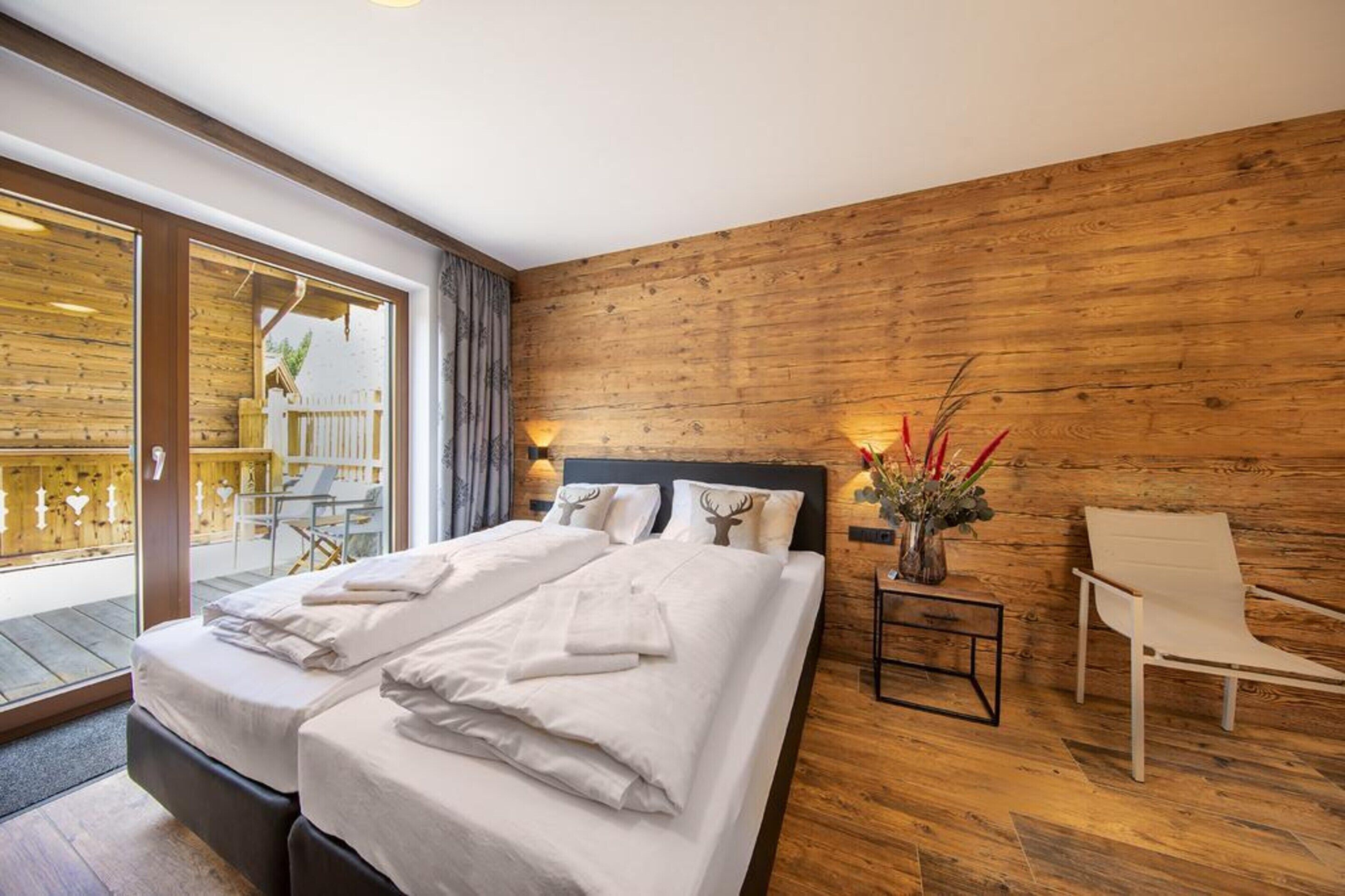 Moderni hotellihuone, jossa on puuseinä, suuri sänky valkoisilla vuodevaatteilla, oleskelualue tuolilla sekä pieni pöytä, jossa on maljakko täynnä kukkia.