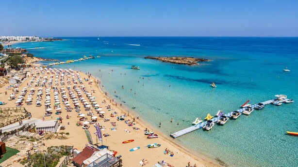 Una playa con filas de tumbonas y sombrillas, un muelle con barcos, y un mar azul cristalino.