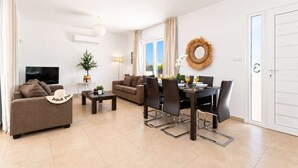Una sala de estar con sofá, mesa de centro, mesa de comedor con sillas, televisor y ventana con cortinas.