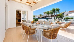 Un espacio de comedor al aire libre cubierto con sillas de mimbre y una mesa, con vista a una piscina y palmeras.