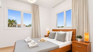 Un dormitorio con cama, dos mesitas de noche, una lámpara y vistas a palmeras a través de las ventanas.