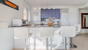 A modern kitchen with white bar stools, a microwave, and a clock on the wall. - Agi Cap Trencat 146 Apartamento for rent in Roses Girona. (Roses)