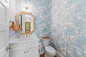 Ein Badezimmer mit weißem Waschtisch, einem Spiegel mit Holzrahmen, einer Toilette und einer Wand mit tropischem Blattmuster.