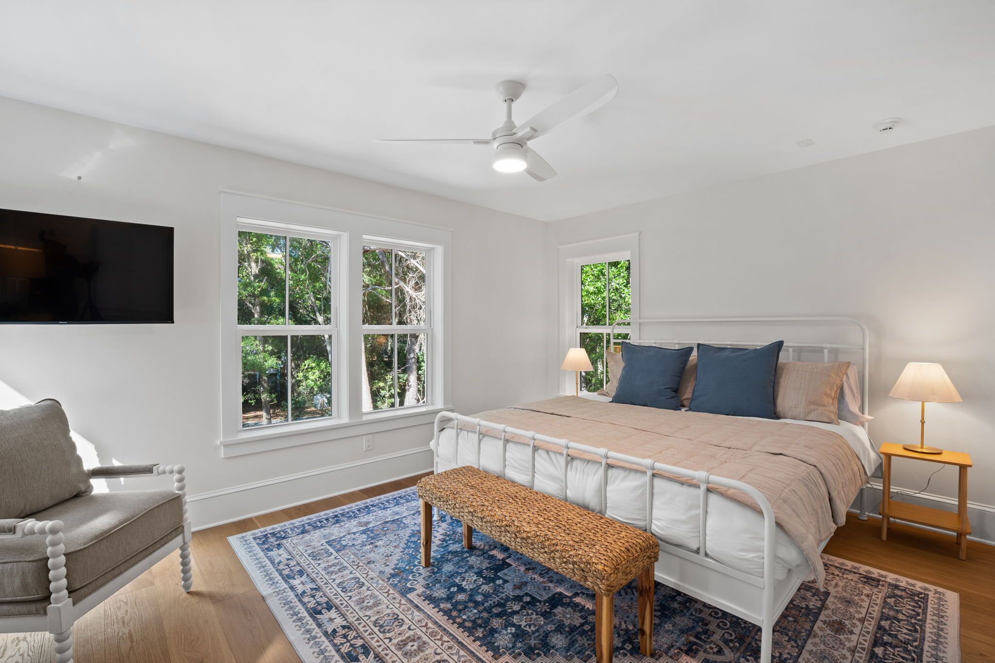 Un dormitorio con cama, banqueta, sillón, televisión y ventana con vista a árboles.