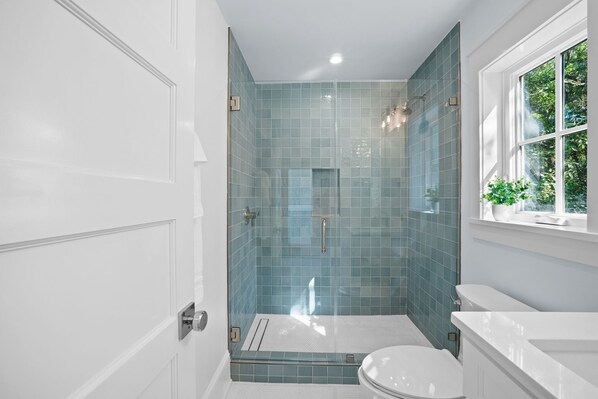 A modern bathroom with a glass-enclosed shower, white fixtures, and a window with a view of greenery.