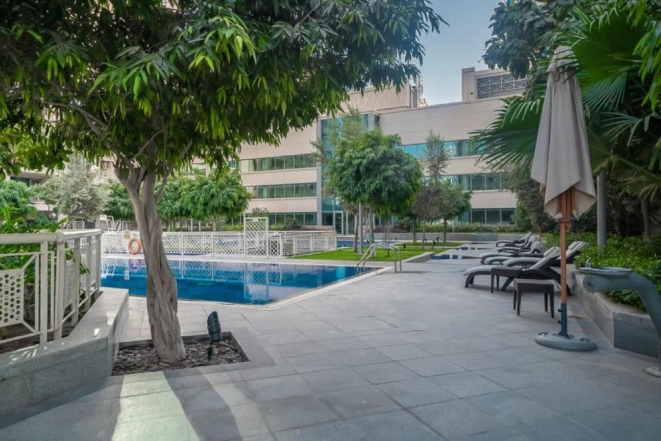 A pool area with lounge chairs and umbrellas, surrounded by greenery and modern buildings.
