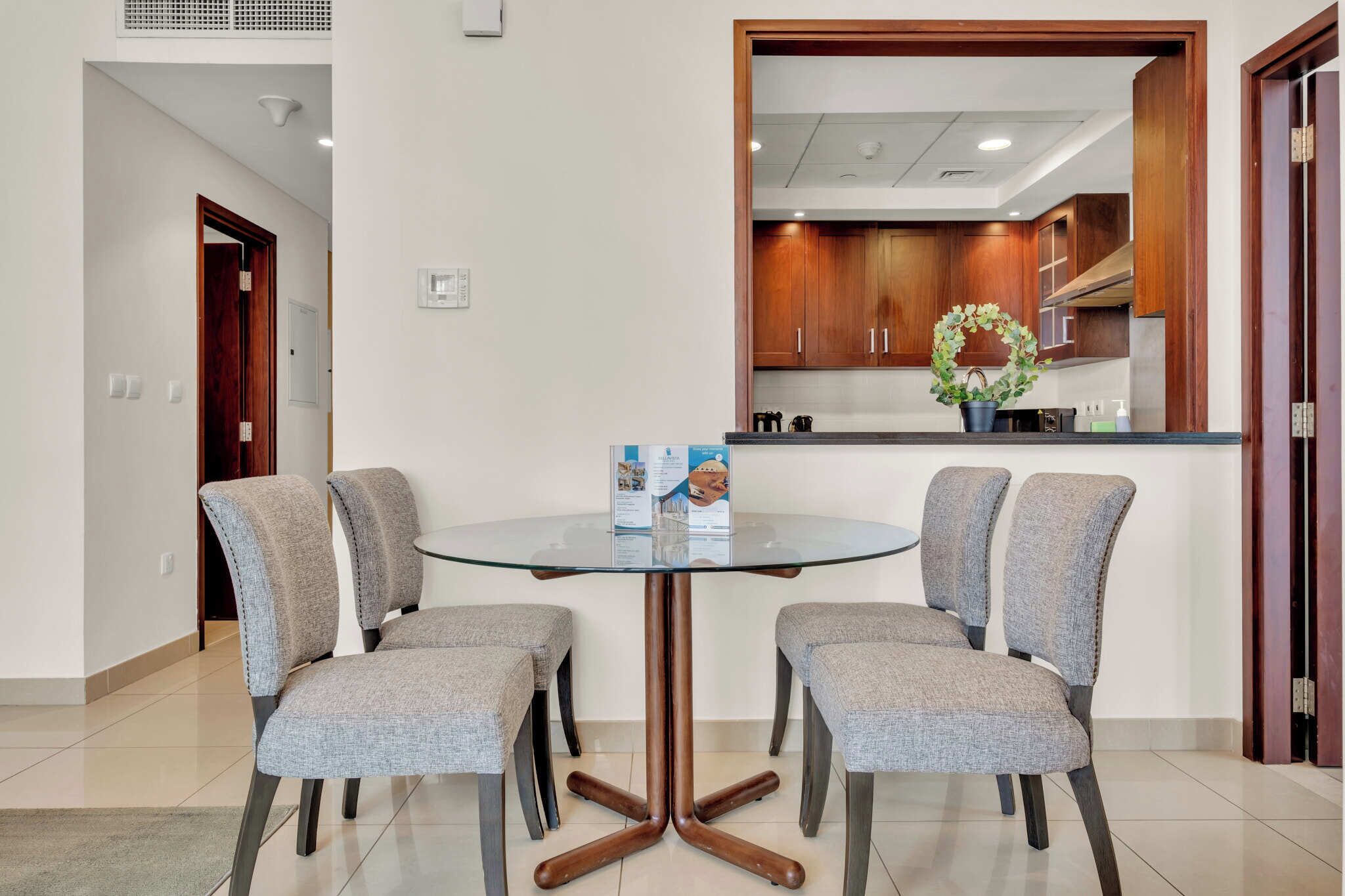 A dining area with a round glass table and four grey upholstered chairs. A brochure is placed on the table.