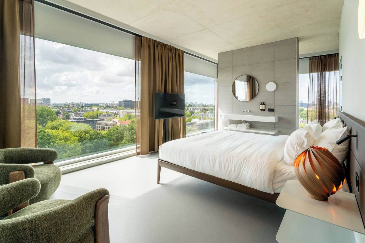 Moderni hotellihuone, jossa on suuri sänky, litteänäyttöinen televisio, pyöreä peili ja suuret ikkunat, joista avautuu näkymä kaupungin ylle.