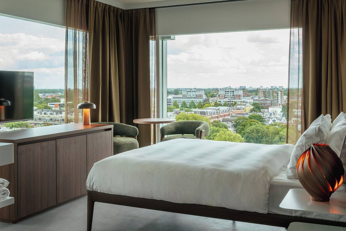 Hotellihuone, jossa on suuri sänky, työpöytä, jossa on televisio, kaksi tuolia, pieni pöytä ja suuret ikkunat, joista avautuu näkymä kaupunkiin.
