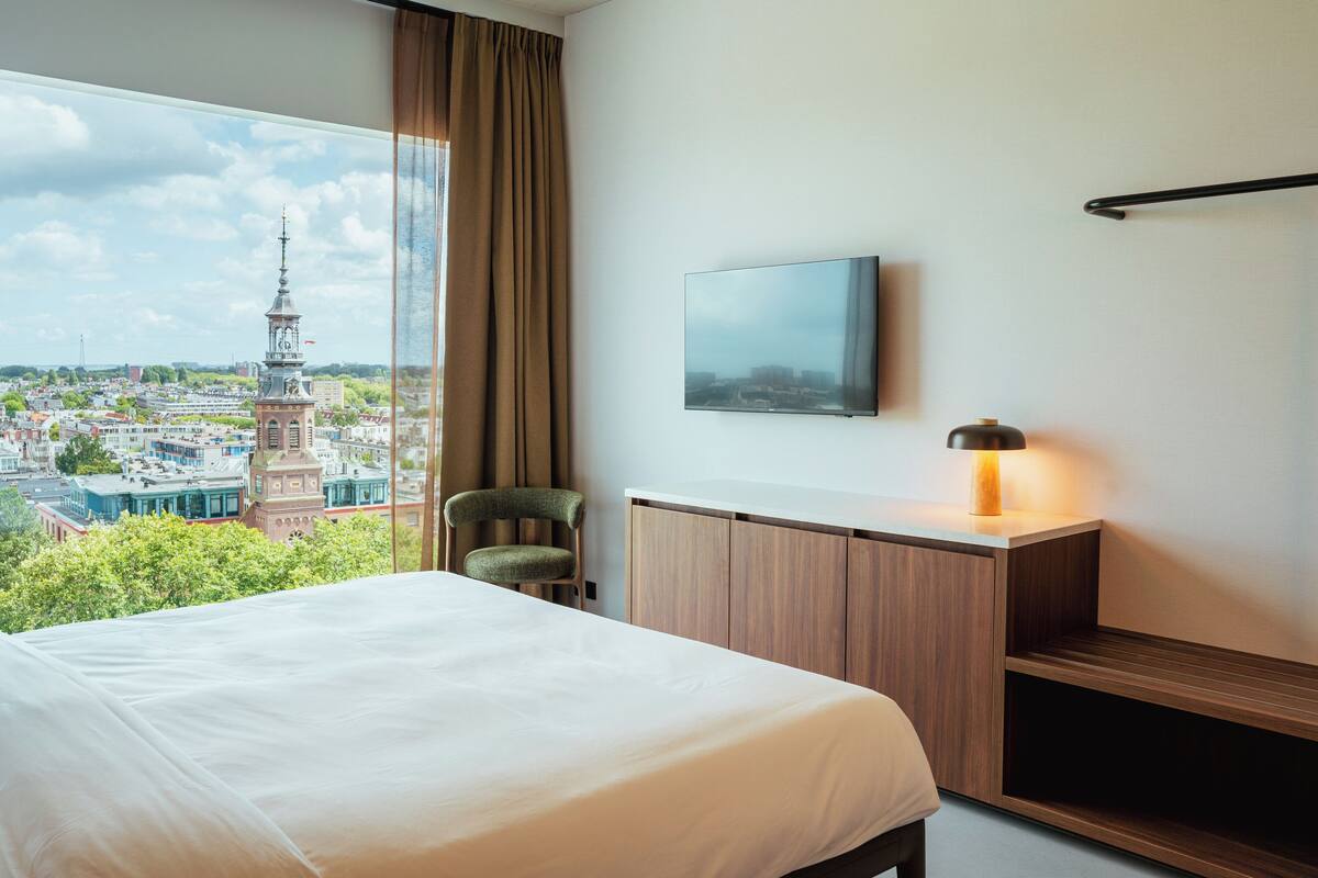 Hotellihuone, jossa on suuri sänky, seinälle asennettu televisio, työpöytä lampun kanssa ja näkymä kaupunkimaisemaan, johon kuuluu myös torni.