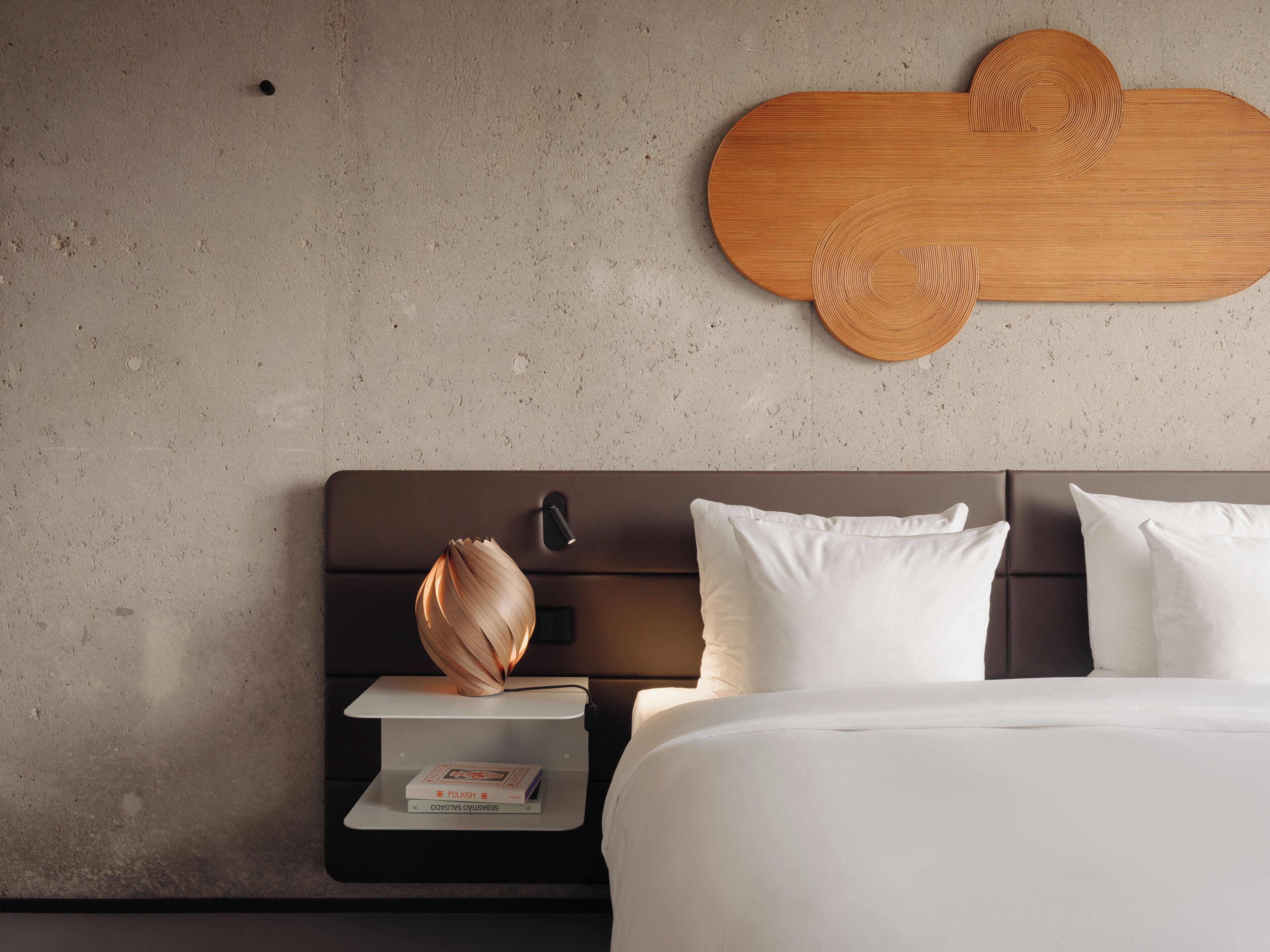 A modern hotel room with a concrete wall, a wooden headboard, a bedside table with a lamp, and a bed with white bedding.