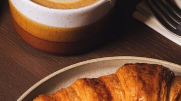Ein Croissant, eine Kaffeetasse mit Latte-Art und eine kleine Schale Obst auf einem Teller.