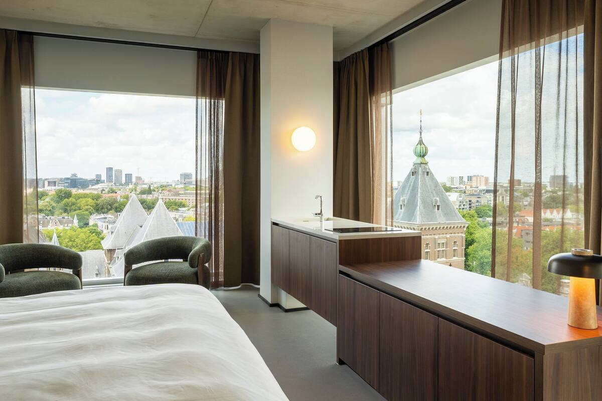Hotellihuone, jossa on sänky, kaksi tuolia, pieni keittokomero ja suuri ikkuna, josta on näkymä kaupunkiin.