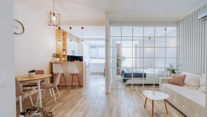 A modern living room with a sofa, a dining area with chairs, a kitchen with a counter, and large windows.