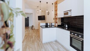 A modern kitchen with white cabinets, a black countertop, and a built-in oven. There are pendant lights, a flat-screen TV, and a small island with a kettle.