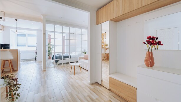 A modern, well-lit living room with a wooden floor, white walls, and a large window.