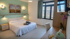 Een slaapkamer met een bed, nachtkastjes, een bureau met stoel, een raam met uitzicht, en een schilderij aan de muur.